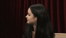 a woman with long black hair is sitting in front of a red curtain and says `` i 'm good '' .