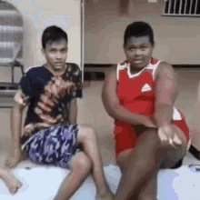 two young men are sitting next to each other on a bed . one of the men is wearing a red adidas jersey .