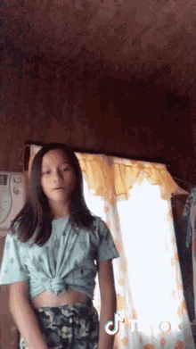 a girl in a crop top is standing in front of a window with a tiktok logo on the bottom right corner .
