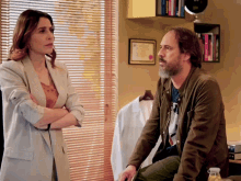 a man and a woman are talking in front of a window with blinds