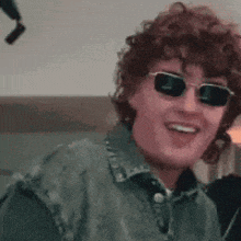 a man wearing sunglasses and a denim jacket is smiling .