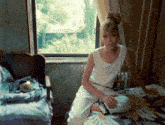 a woman sits on a bed in front of a window and a chair