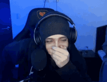 a man wearing headphones and a beanie is sitting in front of a microphone and covering his mouth .