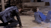 a man in a leather jacket is kneeling next to another man in a blue shirt