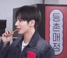 a man in a denim jacket is eating a snack in front of a sign with korean writing
