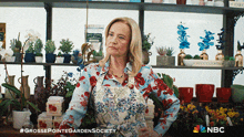 a woman in a floral shirt is standing in front of a shelf full of potted plants and a nbc logo