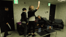 a group of people are standing in a room with a green exit sign on the wall