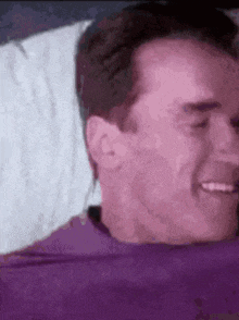 a man in a purple shirt is laying on a bed with a white pillow .