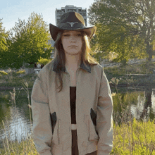 a woman wearing a cowboy hat and a jacket stands in front of a body of water
