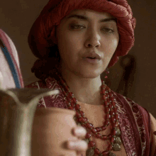 a woman wearing a red turban and a necklace holds a pitcher in her hand
