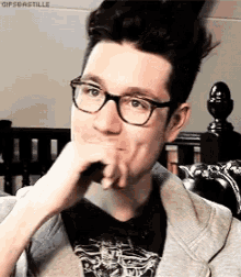 a man wearing glasses and a jacket is sitting on a couch with his hand on his chin .
