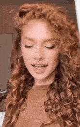 a woman with red curly hair and a nose ring is wearing a brown shirt and earrings .