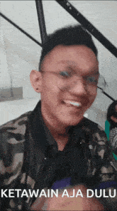 a man wearing glasses and a camouflage jacket smiles for the camera with the words ketawain aja dulu below him