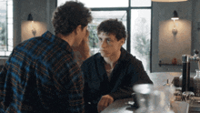 a man in a plaid shirt sits at a bar with another man