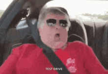 a man wearing sunglasses and a red shirt is sitting in a car with the words test drive above him