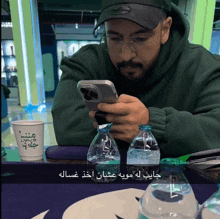 a man sitting at a table looking at his phone next to bottles of water and a cup with arabic writing on it