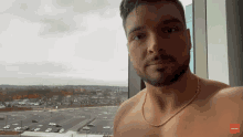 a shirtless man with a gold chain around his neck looks out of a window