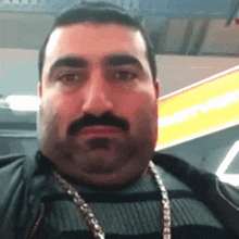 a man with a beard and mustache is wearing a chain around his neck and looking at the camera .
