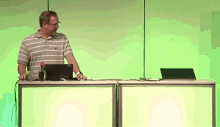 a man in a striped shirt stands at a desk with a laptop and a bottle of water