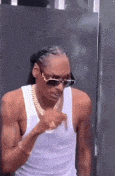 snoop dogg is wearing a white tank top and sunglasses and pointing at something .