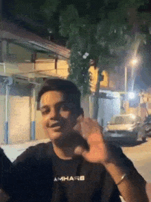 a man wearing a black t-shirt with the word amhare on it is waving his hand in front of a building .