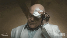 a man in a white suit wipes his forehead with a disney + daredevil logo in the background
