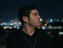 a man in a black hoodie stands in front of a blurry city skyline at night
