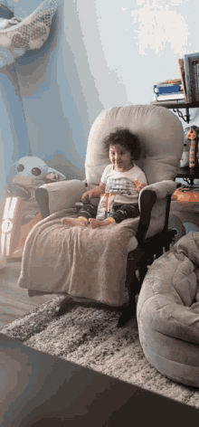 a little boy is sitting in a rocking chair with a blanket on his feet
