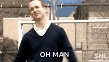 a man in a black sweater is standing in front of a chain link fence and says `` oh man '' .