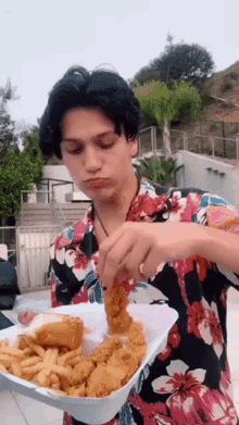 a young man in a floral shirt is eating chicken and french fries from a styrofoam container