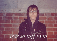 a man in a black jacket stands in front of a brick wall with the words ts is so tuff twin written below him