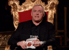 a man in a suit is sitting in a chair with the words here on the stage below him