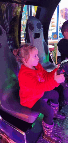 a little girl in a red sweatshirt is sitting in a chair holding a steering wheel