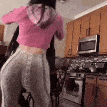 a woman is dancing in a kitchen wearing a pink crop top and gray sweatpants .