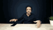 a man in a black shirt is sitting at a desk with his hands outstretched and a phone on the table