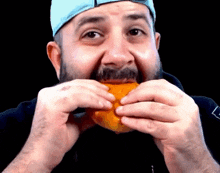 a man with a beard wearing a blue hat is eating a hamburger