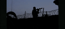 a silhouette of a person standing in front of a fire with a barbed wire fence in the foreground