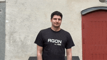 a man wearing a black shirt that says agon by aoc stands in front of a red door