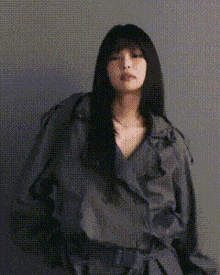 a woman with long black hair is wearing a gray trench coat and standing in front of a wall .