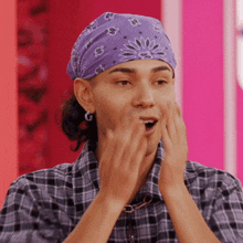 a man wearing a purple bandana and a plaid shirt is covering his face with his hands .