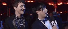 a man in a tuxedo is holding a microphone and talking to another man .