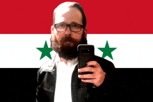 a man with a beard and glasses takes a selfie in front of a syrian flag