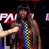 a woman wearing a rainbow checkered dress is being interviewed by a woman holding a microphone that says impact on it