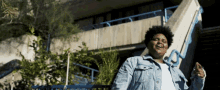 a man in a denim jacket is standing in front of a building and giving a thumbs up