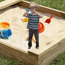 a young boy in a cowboy hat is standing in a sandbox with buckets and shovels