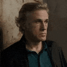 a man in a blue shirt and black jacket is standing in front of a wall and looking at the camera .