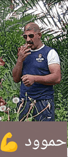 a man wearing sunglasses and a blue shirt smoking a cigarette in front of trees