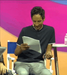 a man in a black shirt is sitting in a chair reading a piece of paper