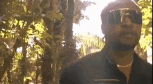 a man wearing sunglasses and a black shirt is standing in a forest .