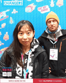 a man and a woman are posing for a photo in front of an olympic channel poster